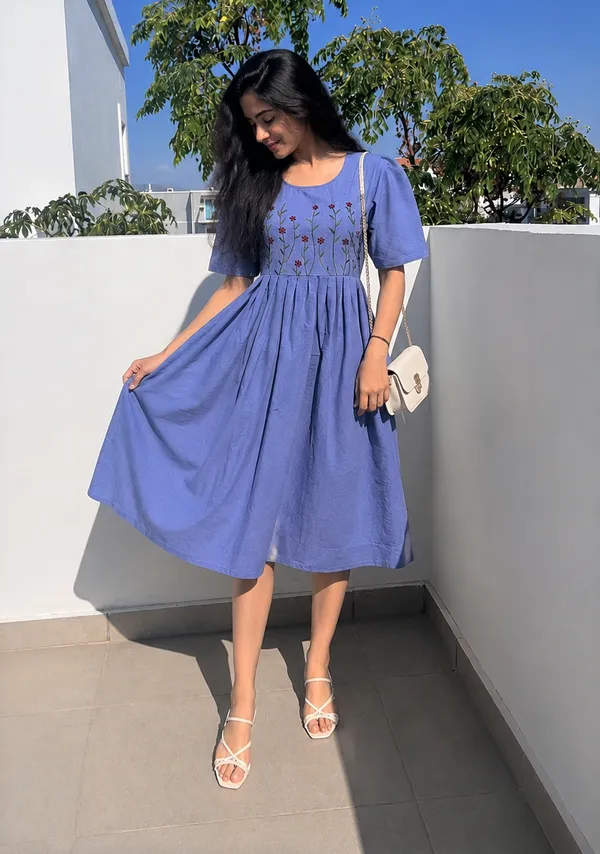 This_is_a_blue_knee-length_A-line_dress_with_short_sleeves_and_a_round_neckline._The_bodice_features_colorful_floral_embroidery._It_is_paired_with_a_beige_shoulder_bag_and_beige_sandals._This_style_is_suitable_for_casual_outings,_brunches,_or_daytime_events.